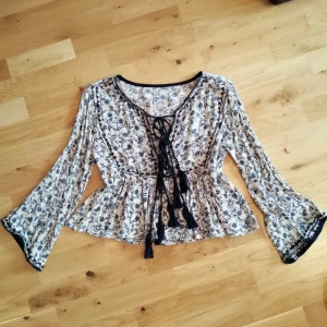 Blommig blus med snörning och vida ärmar - Supersöt blommig blus i vitt och mörkblått med vida ärmar och snygg snörning framtill. Blusen har ett lätt rynkat midjeparti och tunna, flowiga material som ger en bohemvibe. 