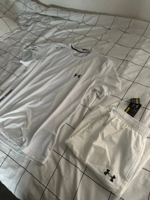 Vitt träningsset från Under Armour - Säljer ett fräscht vitt träningsset från Under Armour med t-shirt och shorts. Båda plaggen har UA-loggan tryckt i svart och är gjorda i ett lätt och snabbtorkande material som passar perfekt till gymmet eller löpning.