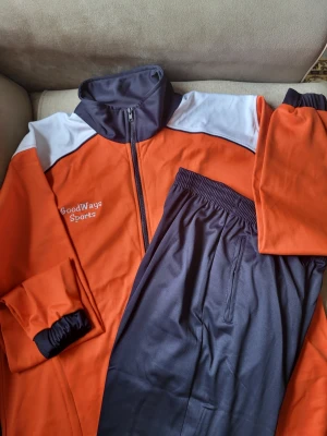 Orange och svart träningskostym GoodWays storlek L bättre XL - Sportig träningskostym från GoodWays Sports i orange och svart med vita detaljer. Jackan har dragkedja och hög krage, byxorna har resår i midjan. Perfekt för dig som gillar en färgstark och bekväm look.