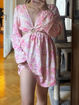 Blommig cut out klänning med ringdetalj - Kort klänning med rosa och vita blommor, djup V-ringning och cut out i midjan med en silverfärgad ringdetalj framtill från New Yorker!
