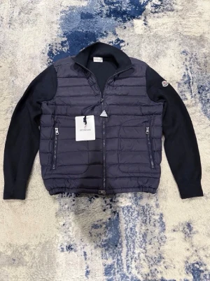 Moncler cardigan mörkblå - Säljer en riktigt snygg Moncler cardigan i mörkblått. Perfekt om du vill ha något som är lite mer dressat än en vanlig dunjacka men fortfarande varmt och bekvämt ✨  • Vadderad front med dun • Stickade ärmar och rygg • Två fickor med dragkedja • Moncler-logga på ärmen • Mörkblå färg  📏 Storlek: (S) ✨ Skick: Mycket bra skick  En av Monclers mest populära modeller och passar till i princip allt 👌  Vid frågor eller om du vill ha fler bilder är det bara att skriva 💌😊