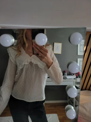 Vit volangblus med prickigt mönster - Söt vit blus med volangdetaljer längs ringningen och diskret prickigt mönster. Blusen har v-ringning och långa, transparenta ärmar i tunn mesh. Perfekt för en trendig och romantisk look. 158/164m använd 1 gång.