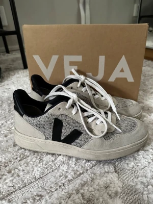Gråa Veja sneakers med svart detalj - Snygga sneakers från Veja i grått och beige med svart V-logga på sidan. Ovandelen är i textil och mocka, med vita skosnören och svart hälparti. Klassisk låg modell som passar till det mesta och ger en clean look.