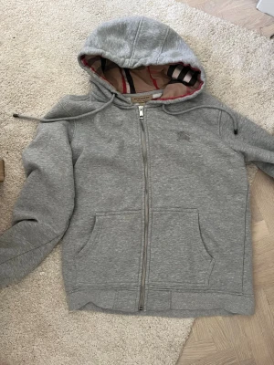 Grå hoodie från Burberry med dragkedja - Snygg grå hoodie från Burberry med dragkedja framtill och klassiskt rutigt foder i huvan. Två stora fickor fram och justerbar dragsko i huvan. Perfekt för en chill och stilren look. Materialet är mjukt och skönt bomullstyg. Helt nytt skick
