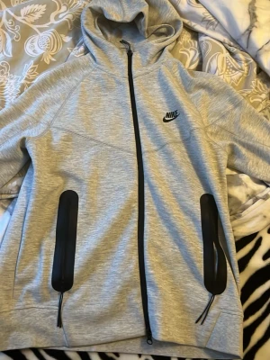 Grå Nike hoodie med dragkedja - Snygg grå hoodie från Nike med svart logga på bröstet. Hoodien har hel dragkedja framtill, två svarta dragkedjeförsedda fickor och en skön huva. Tillverkad i mjukt material som passar perfekt till chill eller träning. Enkel och stilren design som funkar till det mesta.