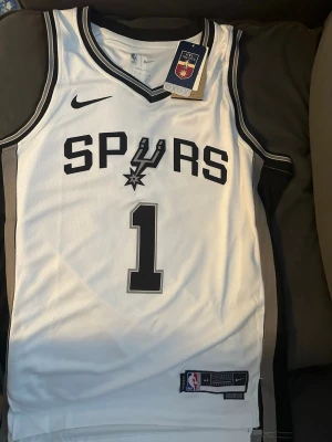 San Antonio Spurs basket tröja wembanyama  - Säljer ett vitt San Antonio Spurs basketlinne från Nike med svart logga och nummer 1 på framsidan. Linnet har svarta och grå detaljer längs sidorna och runt halsen. Perfekt för basketträning eller att bära som streetwear.