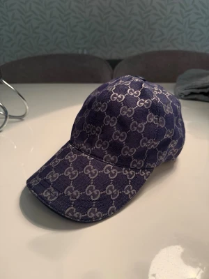 Blå Gucci keps med monogram - Säljer en blå keps från Gucci med det klassiska GG-monogrammet i vitt över hela kepsen. Kepsen har böjd skärm och justerbart spänne bak i läder. Snygg och trendig accessoar som lyfter vilken outfit som helst.