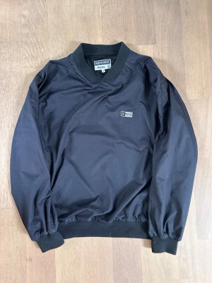 Vintage North West Active Windbreaker Tröja - Svart tröja från NWA med ribbade muddar vid ärmslut och midja. Jackan har en minimalistisk design med liten logotyp på bröstet och rundad krage. Tillverkad i ett lätt, vindtätt material som ger en clean och modern look. Perfekt för vindiga golfrundor. 