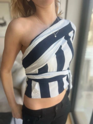Randig oneshoulder topp med knyt - Säljer en snygg randig topp i svart och vitt med oneshoulder-design och knyt i midjan. Toppen har en lång ärm på ena sidan och är ärmlös på andra, vilket ger en unik look. Perfekt för dig som vill sticka ut med en trendig och cool stil.