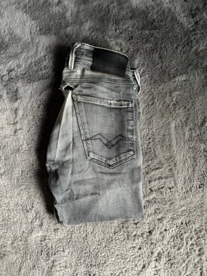 Grå replay anbass jeans - Säljer ett par gråa slim fit jeans från replay med snyggt slitna detaljer och klassisk femficksmodell. Storlek 28/32. Bra skick förutom ett litet hål som syns på bild 4 som lätt borde gå att fixa eller sy ihop. 