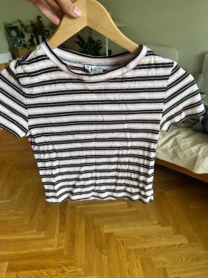 Randig ribbad topp från H&M - Kortärmad topp från H&M med smal passform. Toppen är ribbad och har ett randigt mönster i vitt, svart och rosa. Rund halsringning och stretchigt material som sitter snyggt på kroppen. Perfekt till jeans eller kjol.
