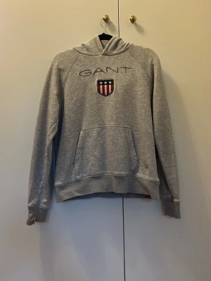 Grå hoodie från GANT med logga - Snygg grå hoodie från GANT med broderad logga och sköld på bröstet. Klassisk känguruficka framtill och mjukt material som känns skönt mot huden. Perfekt för en avslappnad och stilren look. Huva utan dragsko och ribbade muddar vid ärmar och nederkant.