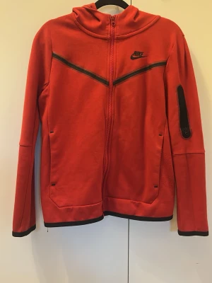 Röd Nike hoodie med dragkedja - Snygg röd hoodie från Nike med hel dragkedja framtill och svarta detaljer. Hoodien har huva, två fickor fram och en extra ficka med dragkedja på ärmen. Tillverkad i mjukt material, perfekt för en sportig och avslappnad stil.
