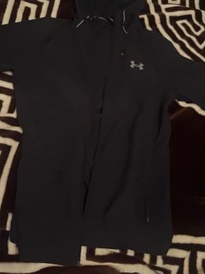 Svart vindjacka från Under Armour - Snygg svart vindjacka från Under Armour med dragkedja framtill och huva. Jackan har en diskret logga på bröstet och praktiska fickor. Perfekt för blåsiga dagar och sportiga outfits. Ord pris 900 används några gånger 