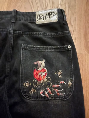 Svarta jeans med broderad björn - Unika svarta jeans från Schyze med en stor broderad björn i röd tröja på bakfickan, omgiven av fiskar och vatten. Jeansen har raka ben och klassisk femficksdesign. Perfekt för dig som vill sticka ut med coola detaljer och streetwear-vibe. Knappen saknas!