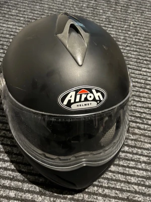 Svart integralhjälm från Airoh - Säljer en svart integralhjälm från Airoh, modell Warrior. Hjälmen har en matt finish, klart visir och ventilationsöppning upptill. Tillverkad i tåligt material och har tydliga logotyper på fram- och sidan. Perfekt för dig som vill ha en stilren och säker hjälm.