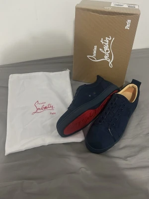 Mörkblå Christian Louboutin sneakers - Snygga mörkblå sneakers från Christian Louboutin med klassisk röd sula och diskret logga på sidan. Skorna är låga och har ovandel i mocka med matchande mörkblå snörning. Perfekta för dig som vill ha en exklusiv och stilren look.