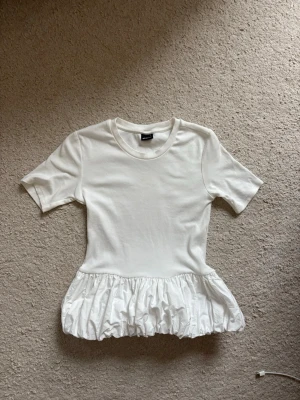 Vit topp med volang från Gina Tricot - Supersöt vit topp från Gina Tricot med korta ärmar och volang nedtill. T-shirten har en enkel rund halsringning och är gjord i mjukt material. Perfekt att styla med jeans eller kjol för en trendig look.