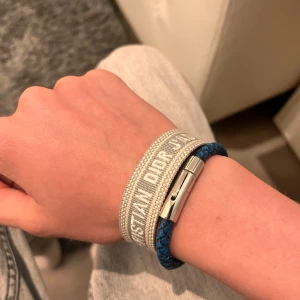 Grått armband från Christian Dior - Säljer ett snyggt grått armband från Christian Dior med broderad logga och tofsar i ändarna. Armbandet har justerbar knytning och är perfekt för att ge din outfit en exklusiv touch. Vet ej om de legit där av billiga priset 