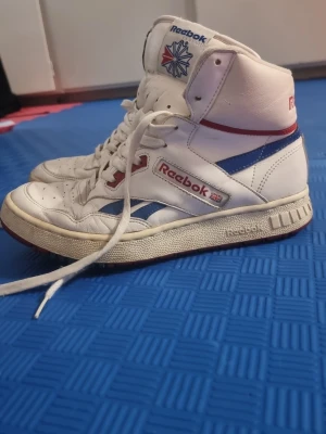 Vita Reebok high top sneakers - Klassiska vita high top sneakers från Reebok med blå och röda detaljer. Skorna har Reebok-logga på sidan och tungan, samt Union Jack-flagga. Tillverkade i läder med perforerade partier för ventilation och kraftig sula. Perfekta för dig som gillar retrostil.