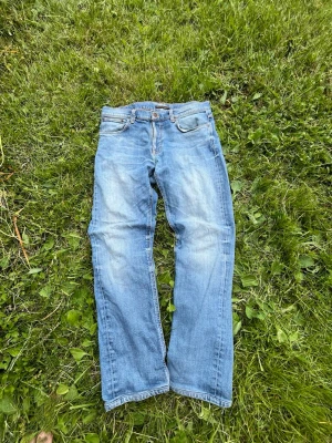 Blå raka slim jeans från Nudie Jeans - Säljer ett par klassiska blå jeans från Nudie Jeans med raka ben och snygg slitning framtill. Jeansen har fem fickor, orange sömmar och är tillverkade i denim. Perfekta för dig som gillar en avslappnad och tidlös stil.