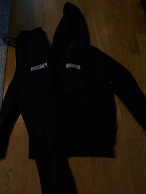 Svart set från Hoodrich med tryck - Snyggt svart set från Hoodrich med matchande hoodie och byxor. Båda plaggen har vitt Hoodrich-tryck på bröstet respektive benet. Hoodien har huva med dragsko och byxorna har resår i midjan. Perfekt för en avslappnad och trendig stil.