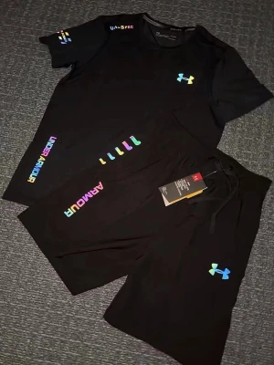 Svart träningsset från Under Armour - Snyggt svart träningsset från Under Armour med färgglada loggor och detaljer. Setet består av en t-shirt och shorts, båda med UA-logga i regnbågsfärger. Perfekt för gymmet eller löprundan. Materialet känns lätt och andas bra. Storlek S, helt ny
