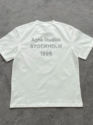 Vit Acne Studios t-shirt Stockholm - Vit t-shirt från Acne Studios med tryck på ryggen där det står 'Acne Studios STOCKHOLM 1996' i grå text. Klassisk passform och rund hals. Perfekt för dig som gillar minimalistisk och stilren design.