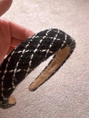 Svart vadderat diadem med rutmönster - Snyggt svart diadem med vadderad form och vitt rutmönster. Utsidan är klädd i ett mjukt tyg som ger en trendig och lyxig känsla. Insidan är beige och har en bekväm passform. Perfekt accessoar för att lyfta din look.