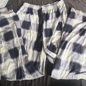 Rutiga pyjamasbyxor med spetsdetalj - Ett par lösa och bekväma pyjamasbyxor med stort rutmönster i svart, vitt och grått. Byxorna har resår i midjan och en dekorativ svart spetsdetalj framtill. Perfekta för mysiga hemmakvällar.