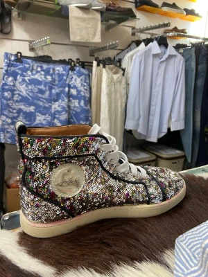 sneakers från Louboutin - Säljer ett par riktigt unika höga sneakers från Christian Louboutin, täckta i färgglada paljetter och med den ikoniska röda sulan. Skorna har vita snören och en tydlig logga på sidan. Perfekta för dig som vill sticka ut och älskar bling!