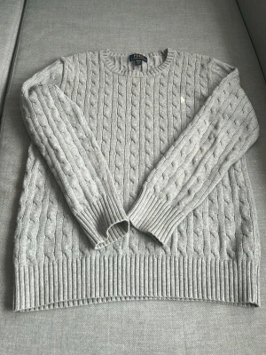 Grå kabelstickad tröja Polo Ralph Lauren - Snygg grå kabelstickad tröja från Polo Ralph Lauren med rund halsringning och diskret broderad logga på bröstet. Tröjan har långa ärmar och ribbade muddar vid ärmslut och nederkant. Perfekt för lager på lager och klassisk stil. Storlek XL (barn), klassas som storlek S. Kostar runt 1000 ny, mitt pris: 300