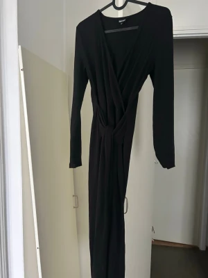 Svart omlottklänning från Bubbleroom - Svart långklänning från Bubbleroom med omlottmodell och knyt i midjan. Klänningen har långa ärmar och är tillverkad i ett mjukt, ribbat material som ger en snygg och bekväm passform. Perfekt för dig som gillar stilrena och tidlösa plagg. Skulle säga att det är s men också m