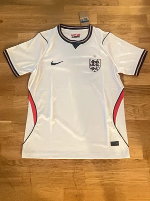 England Nike vit matchtröja - Snygg vit England matchtröja från Nike med broderad landslagssköld och en stjärna ovanför. Tröjan har svarta och röda detaljer längs sidorna och ärmarna, samt rund halsringning med mörkblå kant. Tillverkad i lätt och ventilerande material, perfekt för fotboll.     Finns två styckna i storlek M och en i Storlek L. Fraktar dagen efter elle samma dag som du köpt. 