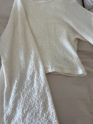 Vit långärmad tröja med vågmönster - Säljer en vit långärmad tröja med unikt vågmönster i tyget. Tröjan har rund halsringning och är tillverkad i ett mjukt, stretchigt material som ger en skön och avslappnad passform. Perfekt för dig som gillar stilrena plagg med en twist.