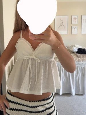 Vit ärmlös linnetopp med knyt - Supersöt vit linnetopp med tunna axelband och volangkant. Toppen har en v-ringning och knyt under bysten som ger en feminin touch. Luftigt material som passar perfekt till sommaren. Perfekt att styla med jeans eller kjol.