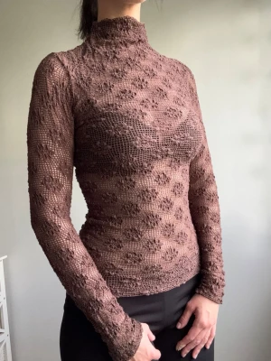 Brun spetsig polotopp med blommönster - Säljer en brun, långärmad polotopp i spets med blommigt mönster. Toppen är tight och har hög hals, vilket ger en elegant och trendig look. Perfekt att styla med både jeans och kjol för en cool vibe.