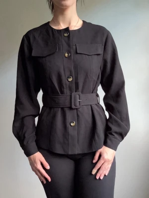 Svart kavaj med bälte och fickor - Stilren svart kavaj med rund halsringning, markerad midja med brett bälte och stora knappar framtill. Två bröstfickor med lock och långa ärmar ger en modern look. Perfekt för dig som gillar minimalistisk och trendig stil.