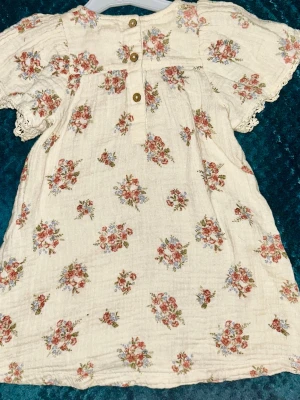 Blommig klänning från Newbie - Söt kortklänning i ljusbeige bomull från Newbie med blommigt mönster i rosa och blått. Klänningen har korta puffärmar med spetsdetaljer vid ärmslut och en lös, luftig passform. Perfekt för sommarens varma dagar.