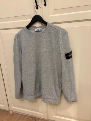 Grå sweatshirt från Stone Island - Snygg grå sweatshirt från Stone Island med klassisk rund hals och ikonisk patch på vänster ärm. Tröjan har ribbade muddar vid ärmslut och nederkant. Perfekt för en avslappnad och stilren look.