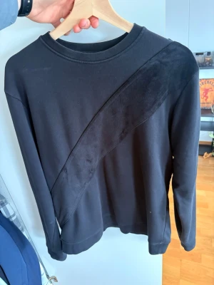 LaVichy tröja - Stilren svart sweatshirt med unik diagonal sammetsdetalj på framsidan. Tröjan har rund halsringning, långa ärmar och knappar på axlarna för en extra touch. Perfekt för dig som gillar minimalistisk design med en twist.