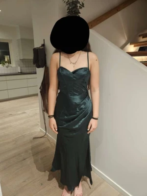 Mörkgrön långklänning med snörning - Elegant mörkgrön långklänning i satinliknande material med smala axelband och v-ringad urringning. Klänningen har öppen rygg med snygg snörning och figurnära passform som ger en lyxig känsla. Perfekt för speciella tillfällen. (Banden vid ryggen ska vara i kors men glömde det när bilden togs) Färgen är mörkgrön men det är svårt att se på bild.