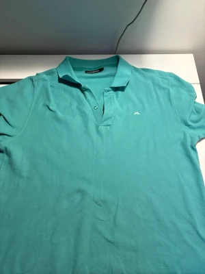 Turkos pikétröja från J.Lindeberg - Snygg turkos pikétröja från J.Lindeberg med klassisk krage och knappslå framtill. Tröjan har en liten vit logga broderad på bröstet och är tillverkad i mjuk bomull som känns skön mot huden. Perfekt för en avslappnad men stilren look.