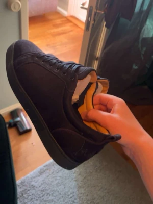 Svarta sneakers från Christian Louboutin - Snygga svarta sneakers från Christian Louboutin med klassisk röd sula och svarta skosnören. Ovandelen är i mocka och insidan har guldfärgade detaljer. Perfekta för dig som vill ha en exklusiv och stilren look. Möts endast upp och tar bara kontanter!