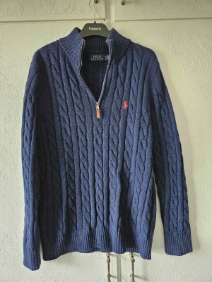 Marinblå kabelstickad tröja Polo Ralph Lauren - Kabelstickad marinblå tröja från Polo Ralph Lauren med halv dragkedja och hög krage. Tröjan har ribbade muddar och nederkant samt den klassiska röda logotypen broderad på bröstet. Perfekt för dig som gillar stilren och tidlös design. (Obs, en R3pl1ka)