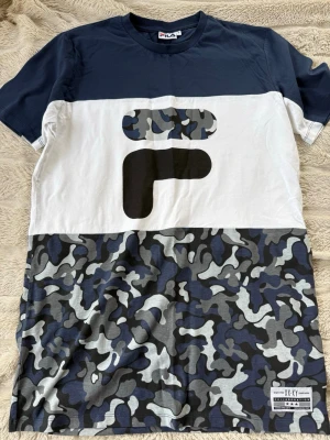 Camouflage t-shirt från FILA - Snygg t-shirt från FILA med marinblå överdel, vit mitt och gråblått camouflage-mönster nedtill. Stor logga framtill och rund halsringning. Perfekt för dig som gillar streetwear och vill sticka ut lite extra.