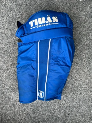 Blå hockeybyxor från Tibås Conovest - Säljer ett par blå hockeybyxor från Tibås Conovest med vita detaljer och logotyp framtill. Byxorna har vadderad insida för extra skydd och komfort, samt justerbart bälte upptill. Perfekta för hockeyträning eller match.