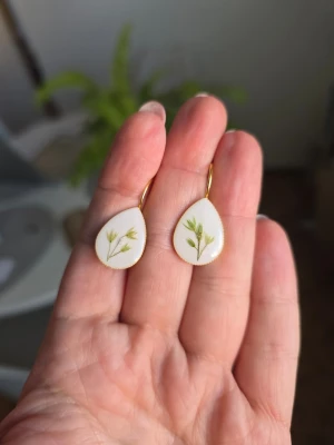Handgjorda örhängen med växt - Unika droppformade örhängen med vit botten och grönt växt inuti. Örhängena har guldfärgade krokar och en stilren, naturlig look. Perfekta för dig som gillar naturinspirerade accessoarer och vill ha något personligt och kreativt. Det finns en defekt i ett örhänge (bubblor)
