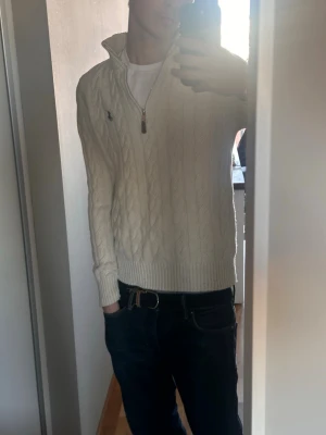 Vit kabelstickad tröja Polo Ralph Lauren - Kabelstickad vit tröja från Polo Ralph Lauren med halv dragkedja och broderad logga på bröstet. Tröjan har ribbade muddar och nederkant, samt klassisk kabelstickad struktur. Perfekt för dig som gillar stilren och tidlös design.