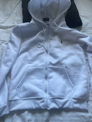 Vit hoodie från Polo Ralph Lauren - Snygg vit hoodie från Polo Ralph Lauren med dragkedja framtill och två stora fickor. Klassisk huva med dragsko och den ikoniska lilla svarta loggan broderad på bröstet. Tillverkad i mjukt bomullsmaterial som är riktigt skönt att ha på sig.